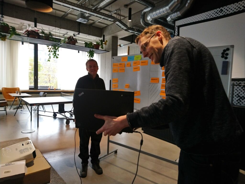 Volker holding a laptop facing away from the camera, towards a whiteboard with sticky notes, Kevin in the background