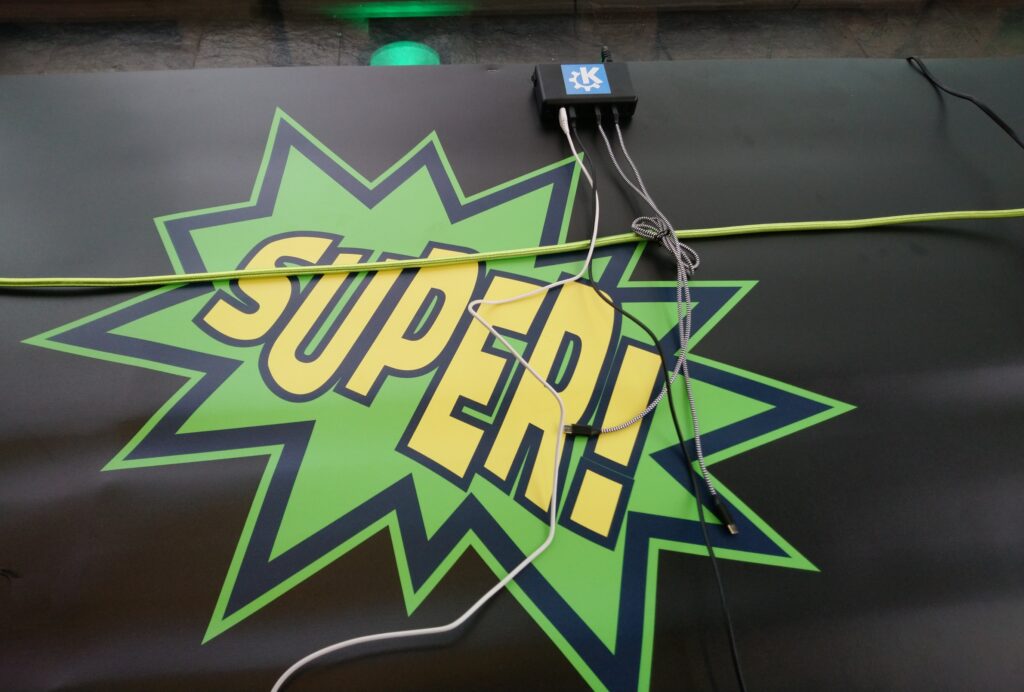 Black cloth covering pallets used as seating accommodation with "SUPER!" written in a comic-like spiky speech bubble, and a KDE-branded USB multi charger in the back
