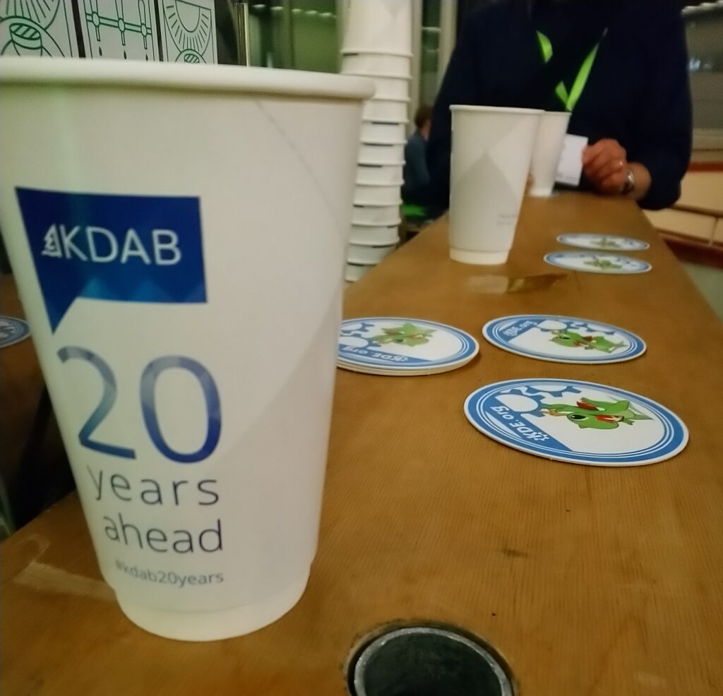 A wooden bar counter with KDAB 20 years ahead cup on it as well as a bunch of KDE-branded beer coasters with Konqi on it