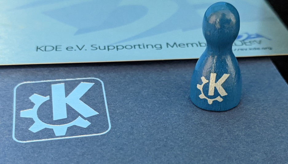 A blue game piece with KDE logo sitting ontop of a blue cardboard box with KDE logo on it and behind it a silver credit card sized sheet reading "KDE e.V. Supporting Member"