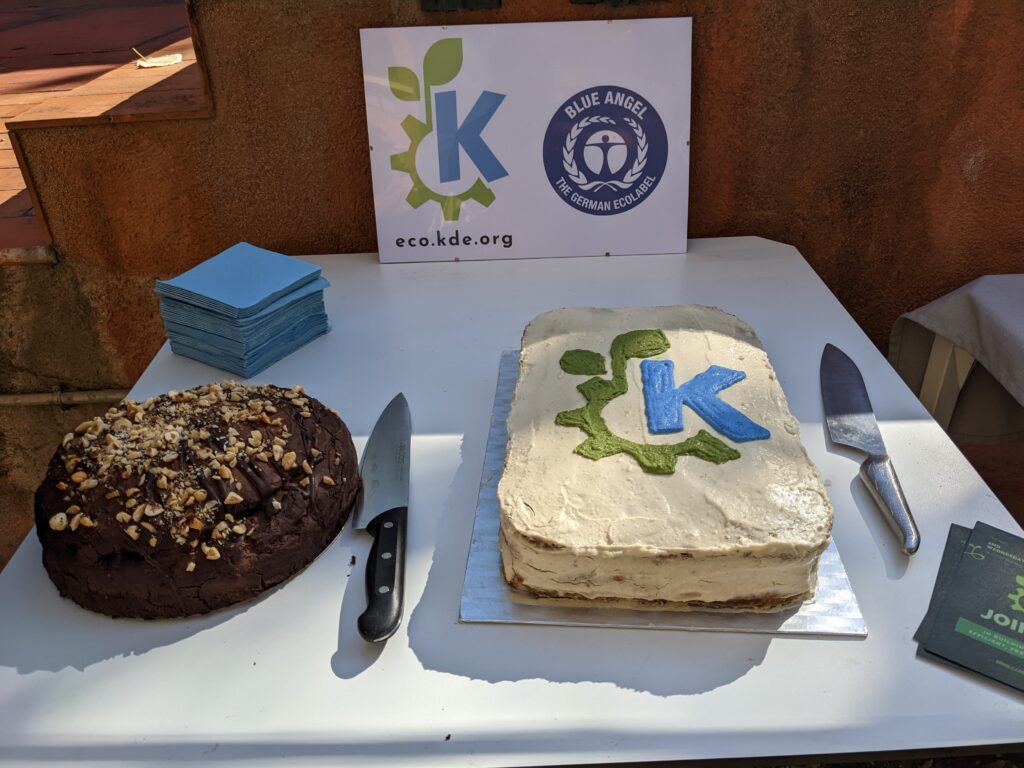 A table with a nut-cream cake with the KDE Eco logo on it, the KDE Eco logo and Blue angel logo on a display behind it