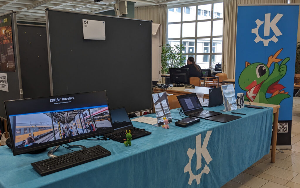 KDE booth at LinuxDay, a table with blue table cloth, KDE logo printed on it, KDE banner in the background. Various monitors, devices, and input devices on the table.