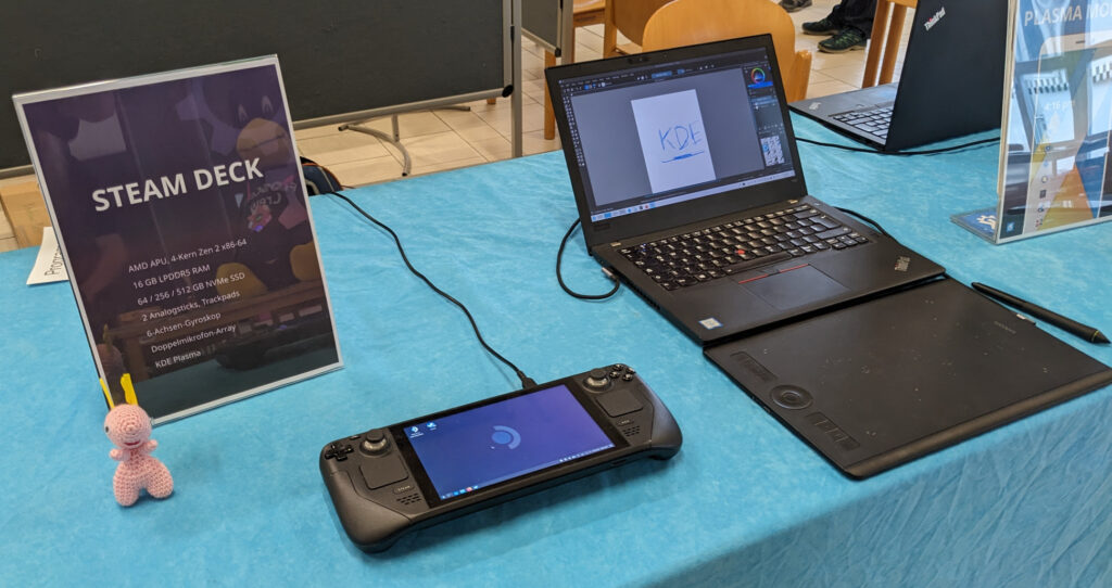 Close-up of booth table, Steam Deck (a handheld gaming console) on the left, “guarded” by a plush Katie (Konqi’s girlfriend). To the right a laptop running Krita with a graphics tablet in front.