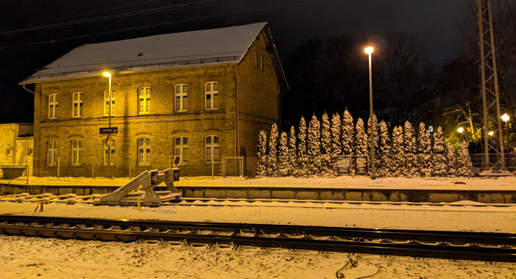 Night, harsh yellow sodium vapor lighting. Train station platform covered in snow, buffer stop in the foreground, station building in the background.