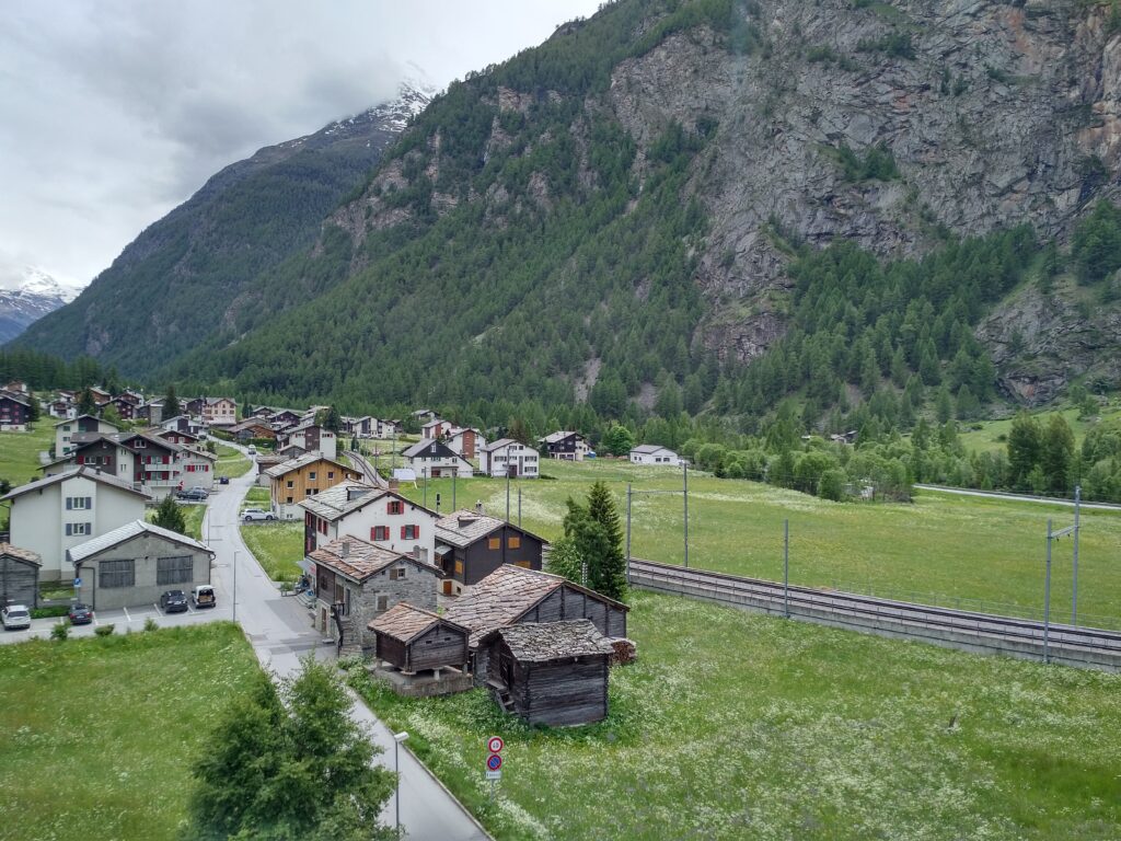 Picture of Randa vaillage, a single road with houses on either side, railway tracks to the right, rest mostly green lawns. Background shows mountains.